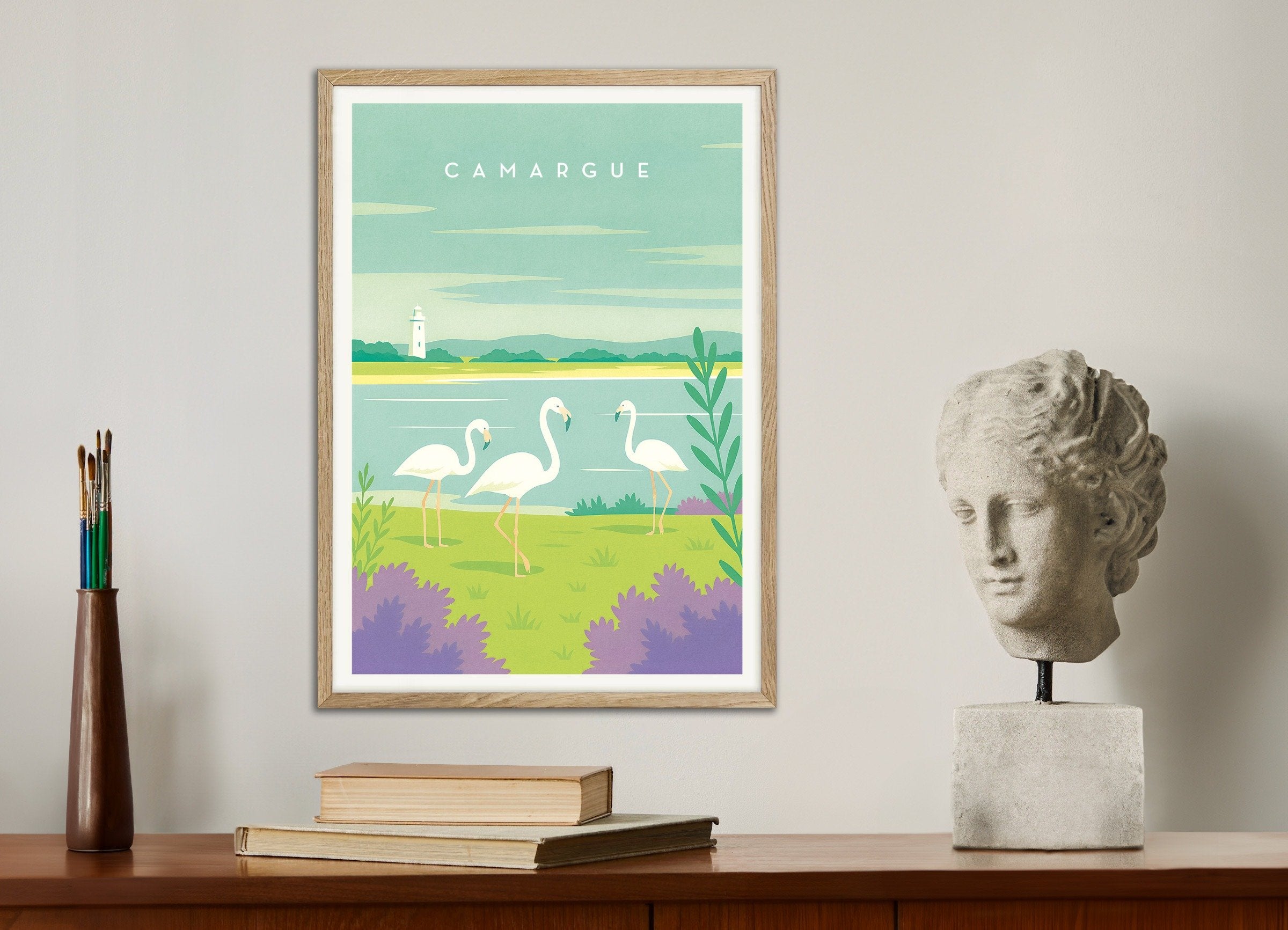 Framed artwork of flamingos in a natural setting on a wall above a wooden shelf with books and a sculpture.