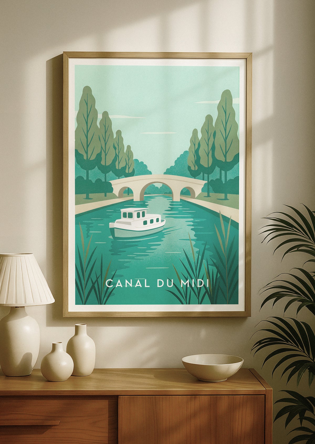 En indrammet rejseplakat med Canal du Midi i Frankrig, med en retrobåd på turkis vand, træer og en stenbro. Den hænger på en beige væg over et konsolbord af træ med dekorative vaser og en lampe.